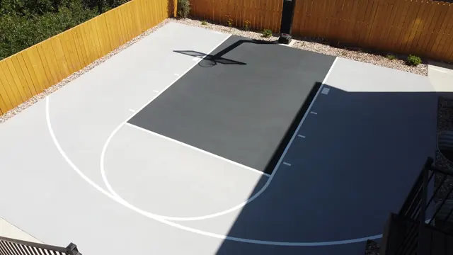 Gray backyard basketball half court with a dark key, white lines and wood perimeter fencing.