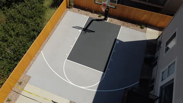 Gray backyard basketball half court with a dark key, white lines and wood perimeter fencing.