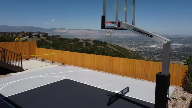 Gray backyard basketball half court with a dark key, white lines and wood perimeter fencing.