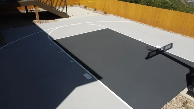 Gray backyard basketball half court with a dark key, white lines and wood perimeter fencing.