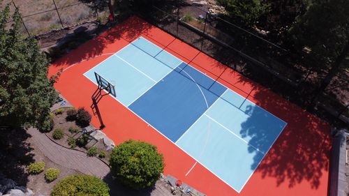 A vibrant outdoor basketball court with blue and red paint surrounded by trees and landscaping.
