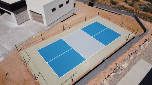 A newly constructed pickleball court with blue playing areas and beige borders next to a house.