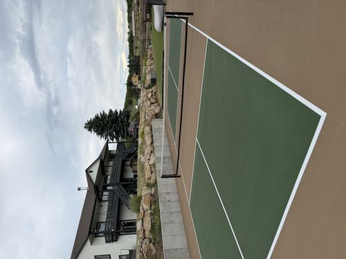 Green outdoor pickleball court with a tan perimeter and center net, beside a stone retaining wall and home.