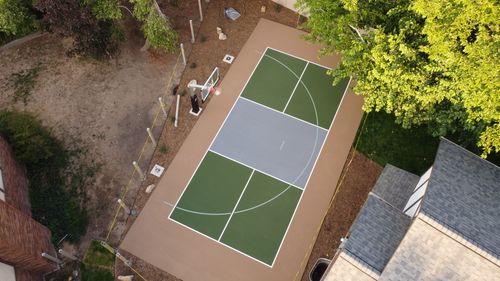 Aerial view of a backyard basketball court next to a house and trees with a basketball hoop on one end.