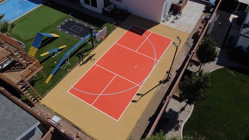 Outdoor basketball court with red and tan surface, adjacent to a backyard with play equipment and putting green.