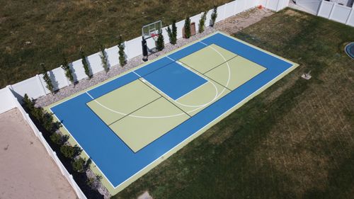 A backyard basketball court with blue and green markings and a single hoop near a white fence.