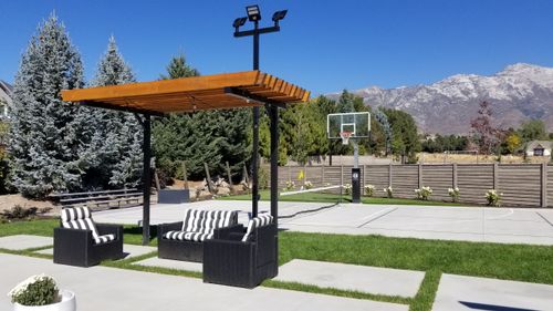 Outdoor seating area with striped cushions under a wooden pergola beside a basketball court and mountains.
