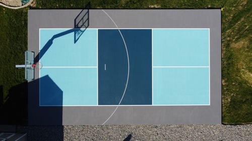 Aerial view of a clean outdoor basketball court with blue and gray sections and a hoop casting a shadow.