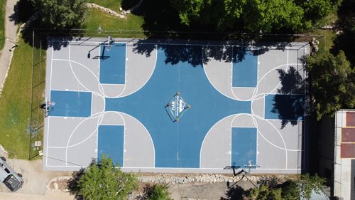 Aerial view of an outdoor basketball court with blue and gray paint and four hoops, surrounded by trees.