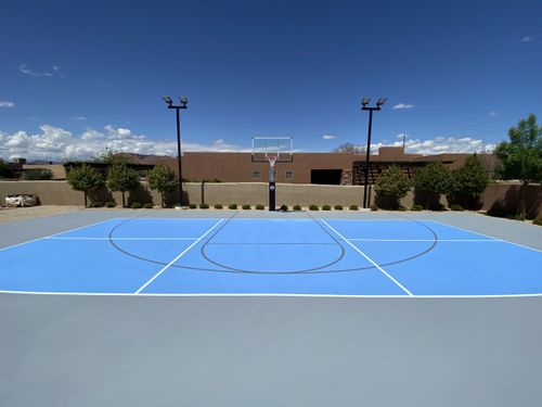 Outdoor basketball court with blue playing surface, hoop, and clear sky background.