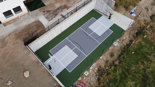 Aerial view of an outdoor basketball court with green and gray surface beside a building and dirt area.
