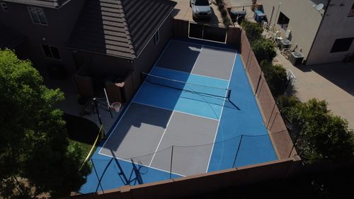 Outdoor multi-sport court with blue and gray zones, a basketball hoop, and a tennis net next to houses.