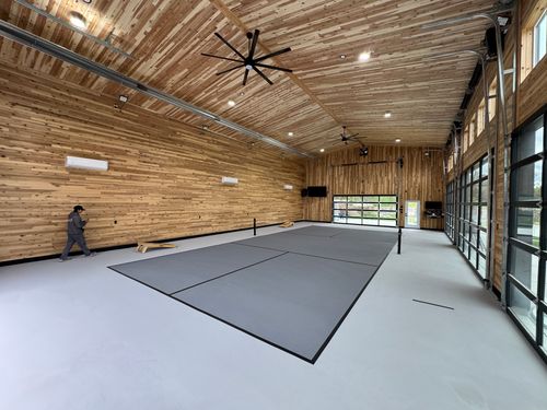 Indoor sports court with gray playing area, wood-paneled walls, ceiling fans, and glass garage doors.