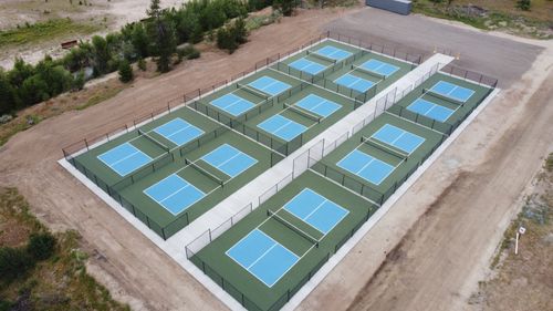 Aerial view of ten blue and green pickleball courts with black fencing on a dirt site.