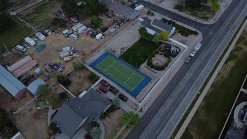 Aerial view of a fenced tennis court beside houses, a playground, vehicles, and a street.