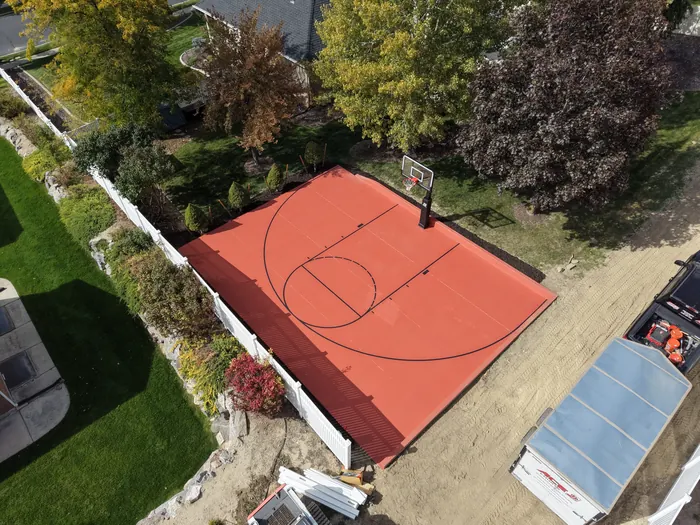 Overhead angle of the red basketball court beside mature trees and a white fence