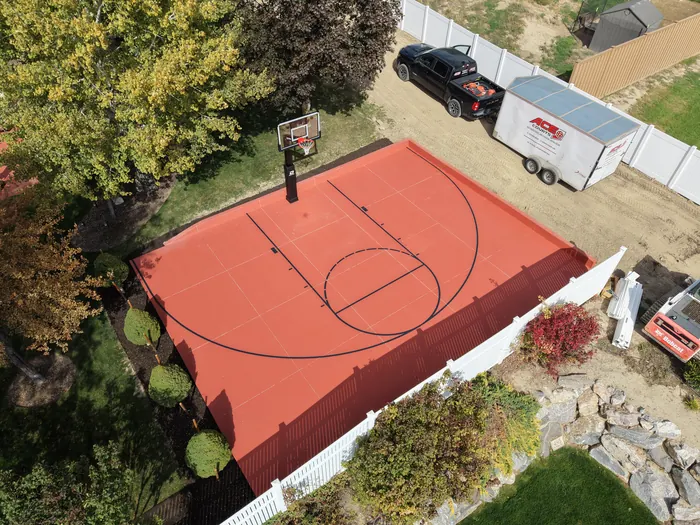 Red backyard basketball court with a black hoop and surrounding fence viewed from above