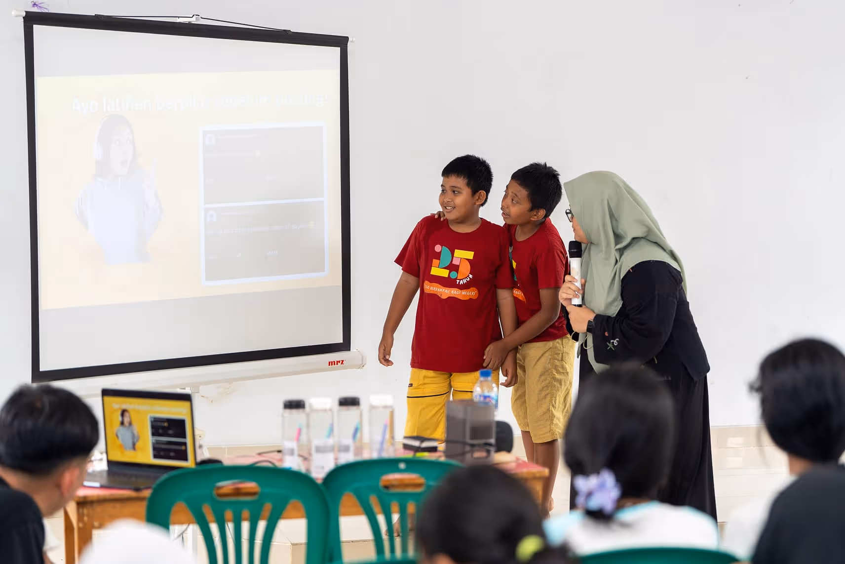 Two boys in matching red shirts stand next to a woman in a hijab holding a microphone, presenting in front of a projector screen to an audience.