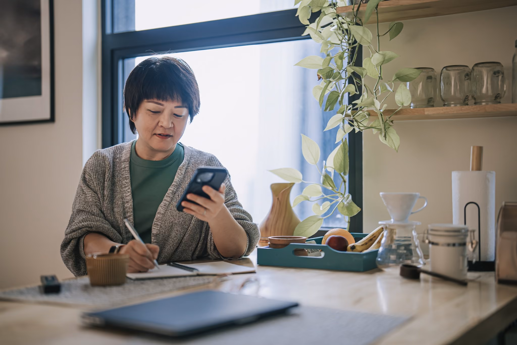 Woman reading a message on her phone and writing down notes at her kitchen table