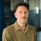 Portrait of a man with short dark hair and a mustache wearing an olive-green collared shirt, seated in an indoor setting with a blurred background.
