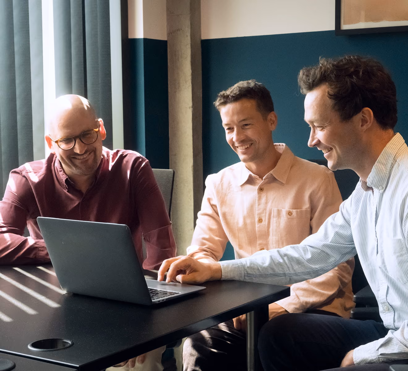 Three men sitting at a table, smiling and looking at a laptop screen in a modern office with teal and white walls and vertical blinds.