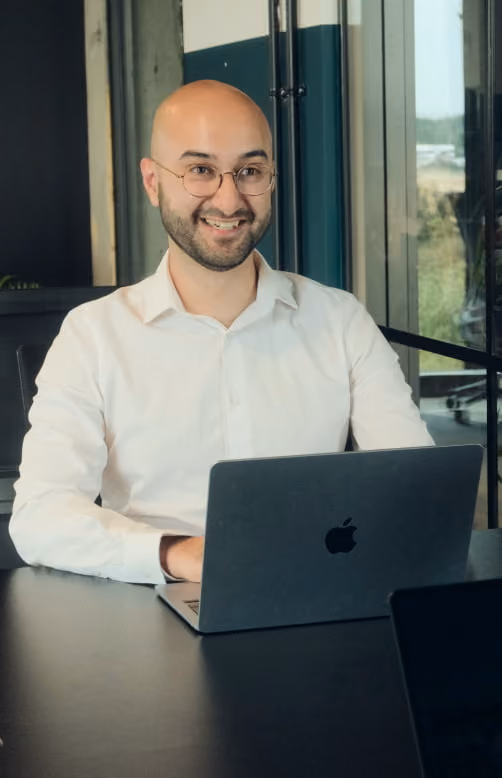 Smiling bald man with glasses wearing a white shirt sitting at a table using a MacBook laptop in a modern office.