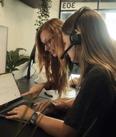 Two women collaborating at a desk with a laptop; one woman wearing glasses points at the screen while the other wears a headset and types.