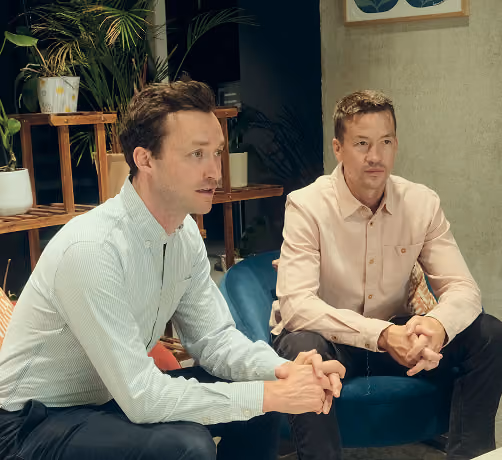 Two men sitting in a room with indoor plants and modern furniture, engaged in a conversation or listening attentively.