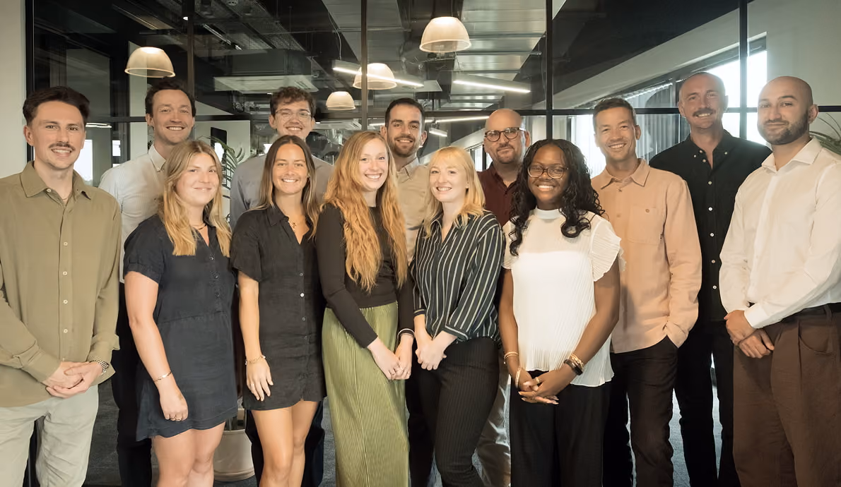 Group of twelve diverse people standing closely together in an office with exposed ceilings and large windows, smiling at the camera.