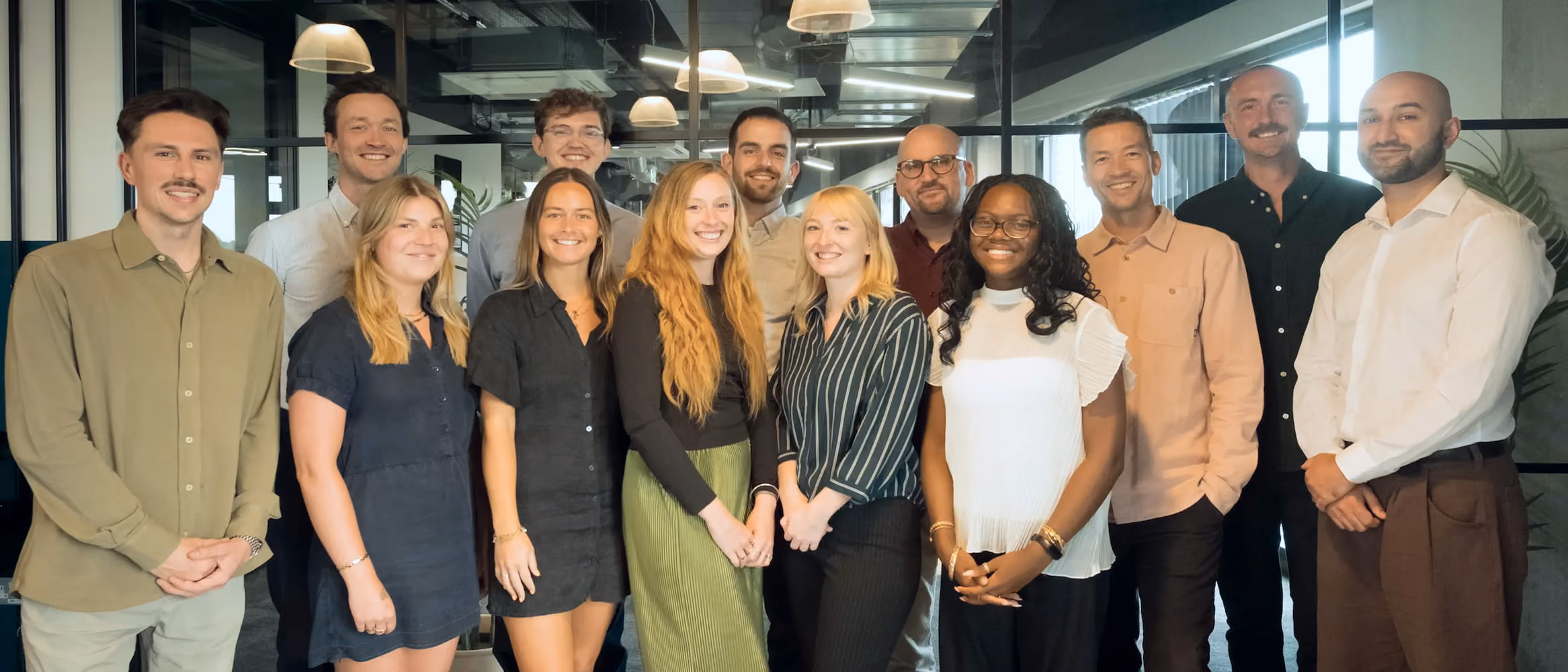 A diverse group of 13 smiling professionals standing indoors in an office space with modern lighting and large windows in the background.