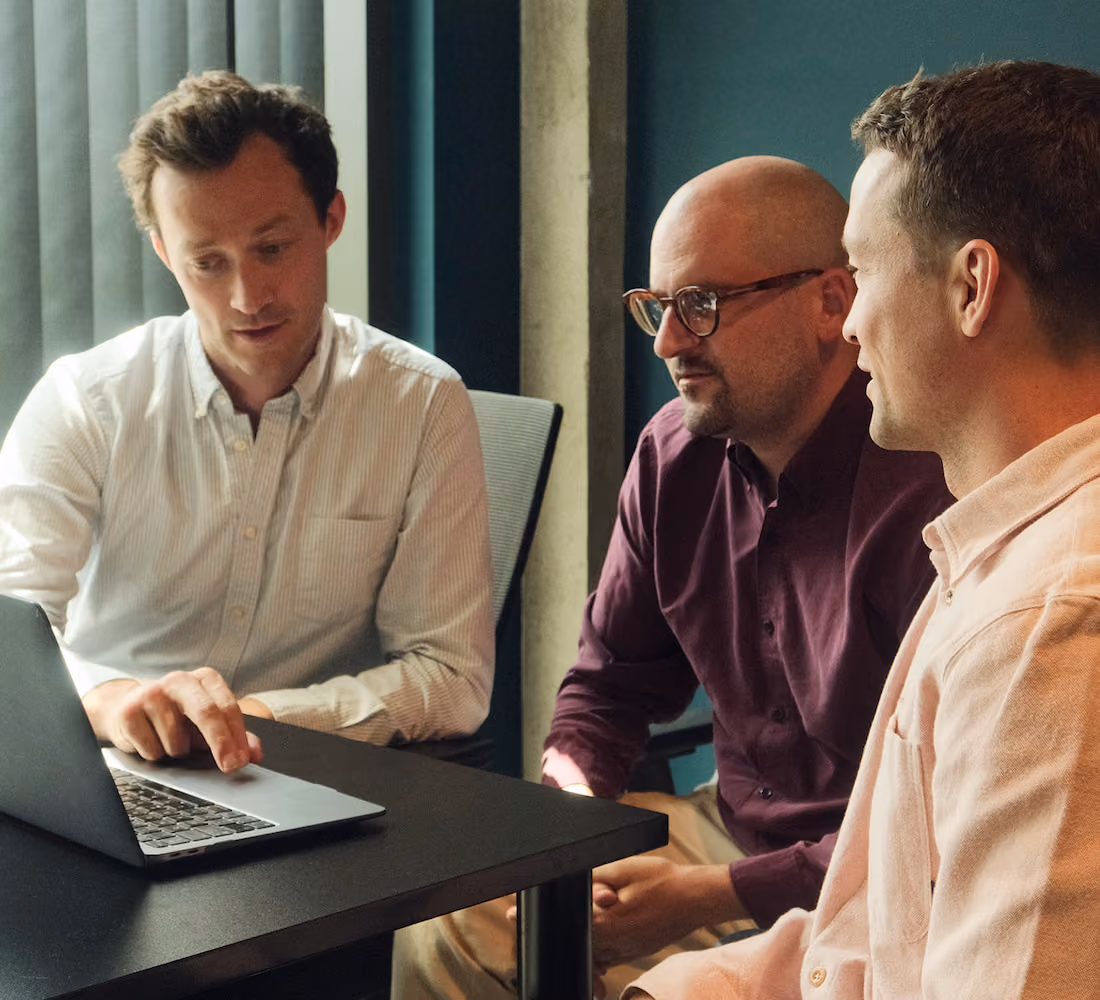 Three men sitting at a black table in an office setting, one man wearing a white shirt is pointing at a laptop screen while the other two men watch attentively.