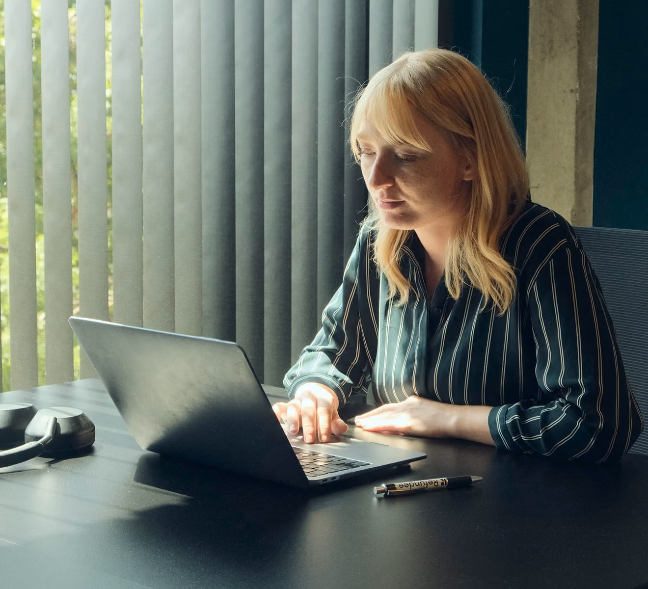A woman with blonde hair wearing a striped shirt sits at a black table typing on a laptop, with vertical blinds and natural light in the background.