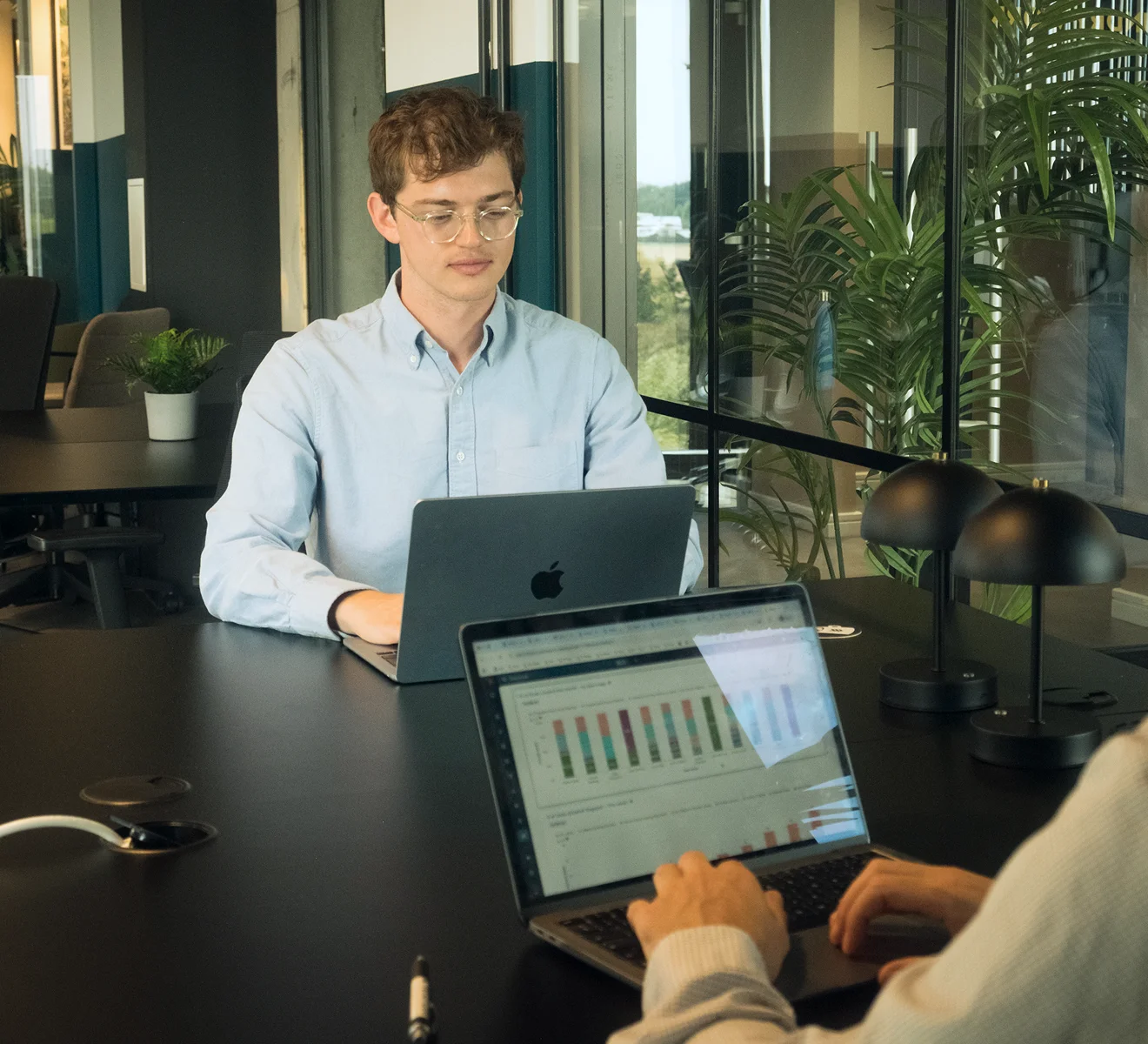 Two people working on laptops in a modern office with glass walls and green plants, one person in a light blue shirt facing forward and another in white using a laptop displaying bar charts.