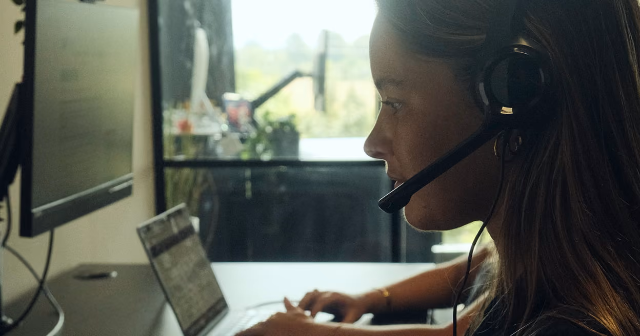 Profile of a woman wearing a headset, working on a laptop at a desk with a monitor, in a softly lit room with a window showing greenery outside.
