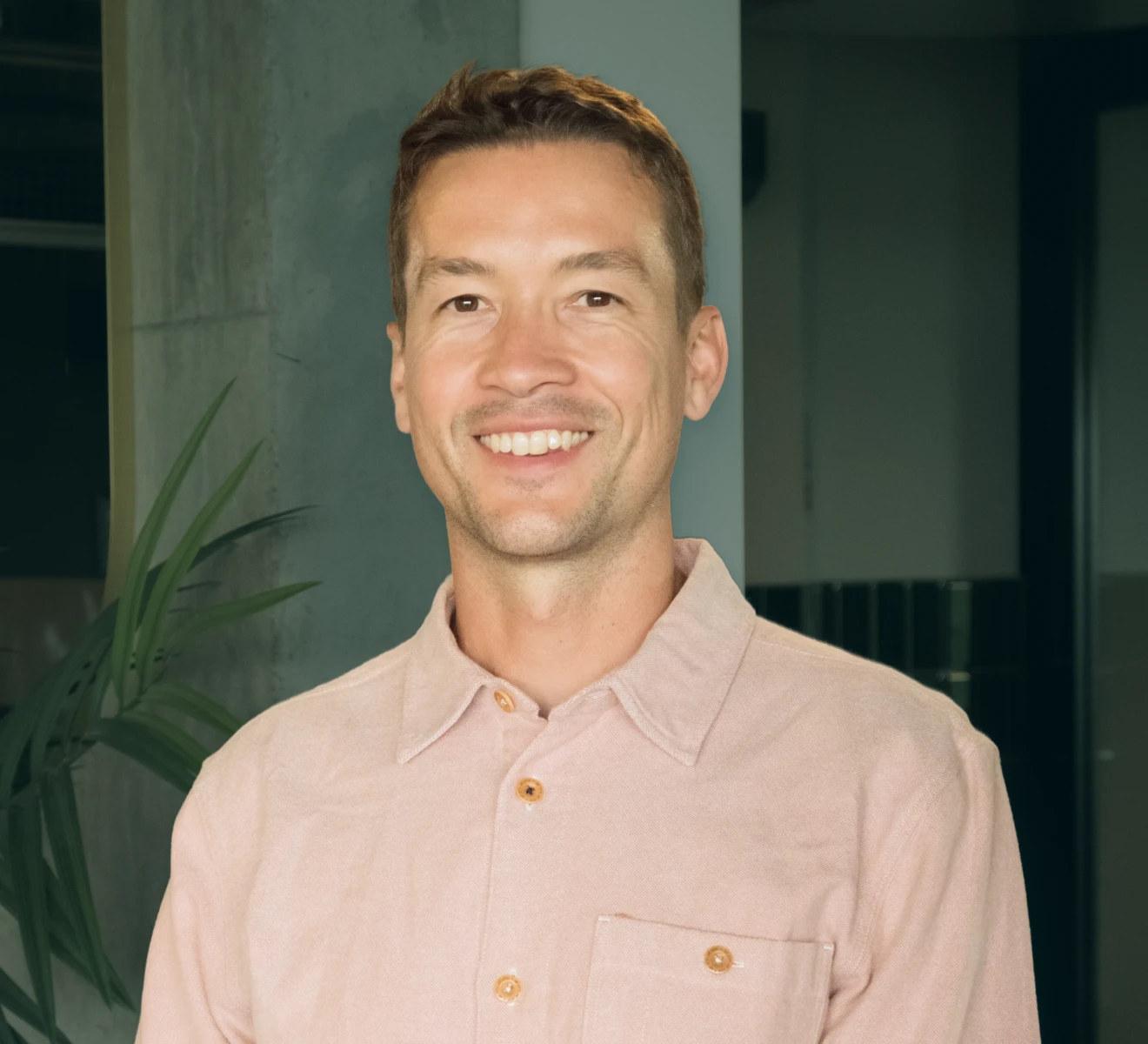 Smiling man with short brown hair wearing a light pink button-up shirt, standing indoors near a concrete wall and a green plant.