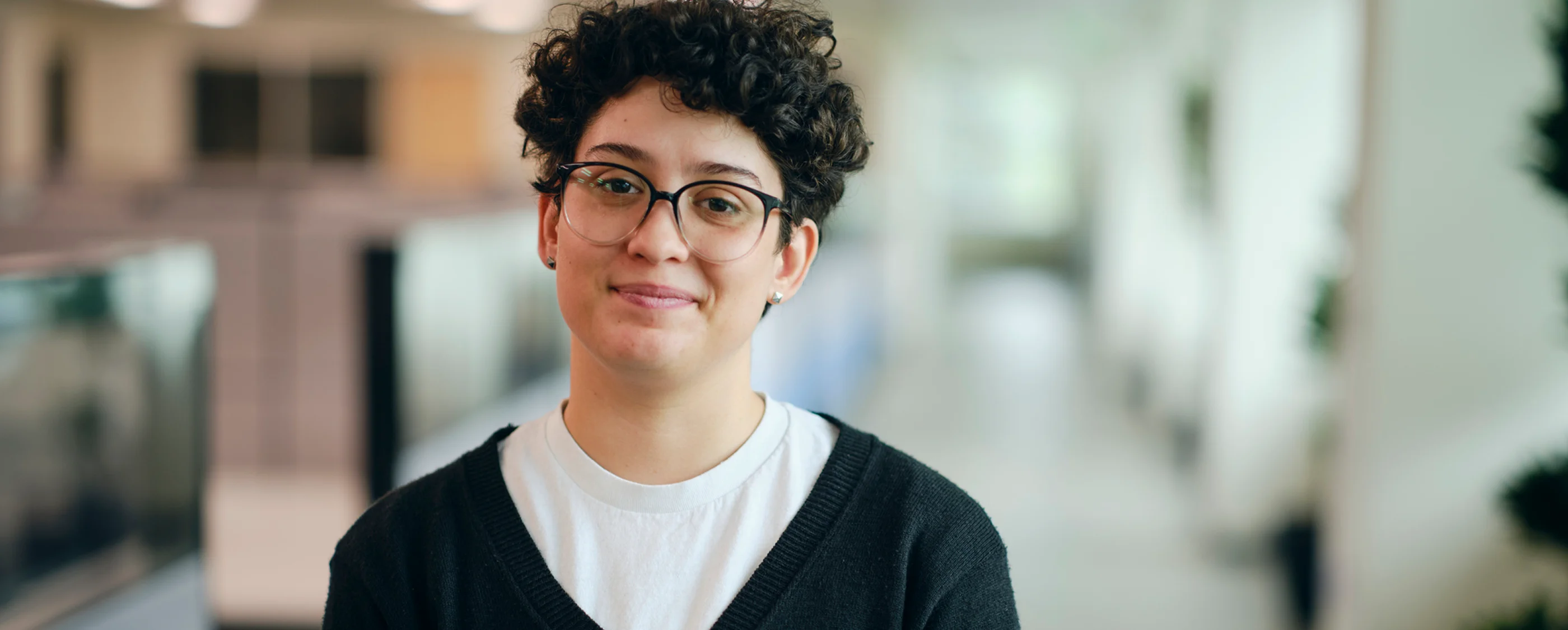 Smiling person with short curly hair, glasses, white shirt, and black sweater standing indoors in a softly blurred modern environment.
