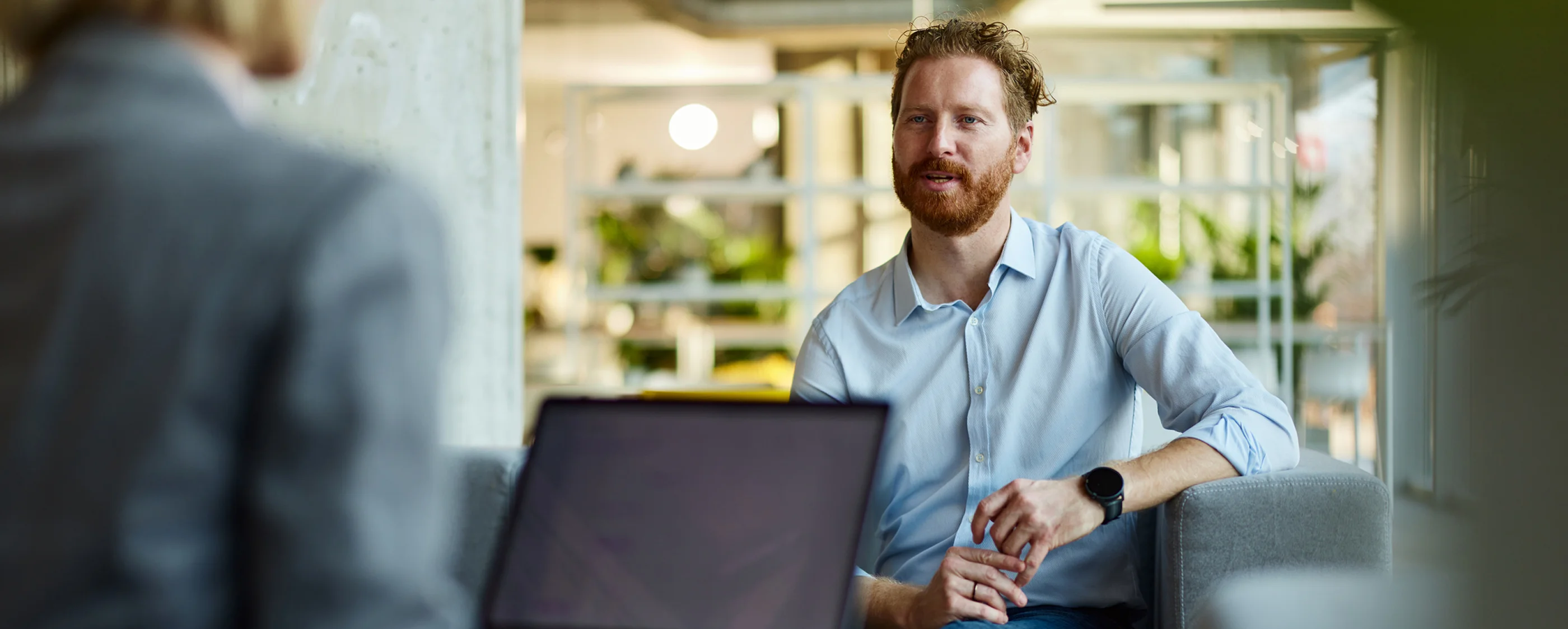 A man with red hair and beard wearing a light blue shirt and smartwatch is seated on a gray couch, talking to a person in a gray suit with a laptop in the foreground, in a bright modern office space with plants and shelves in the background.