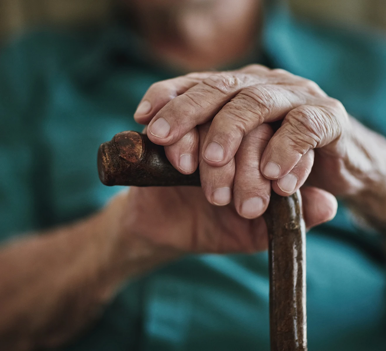 Close-up of elderly hands resting on the handle of a wooden cane, with a blurred background showing a person in a teal shirt.