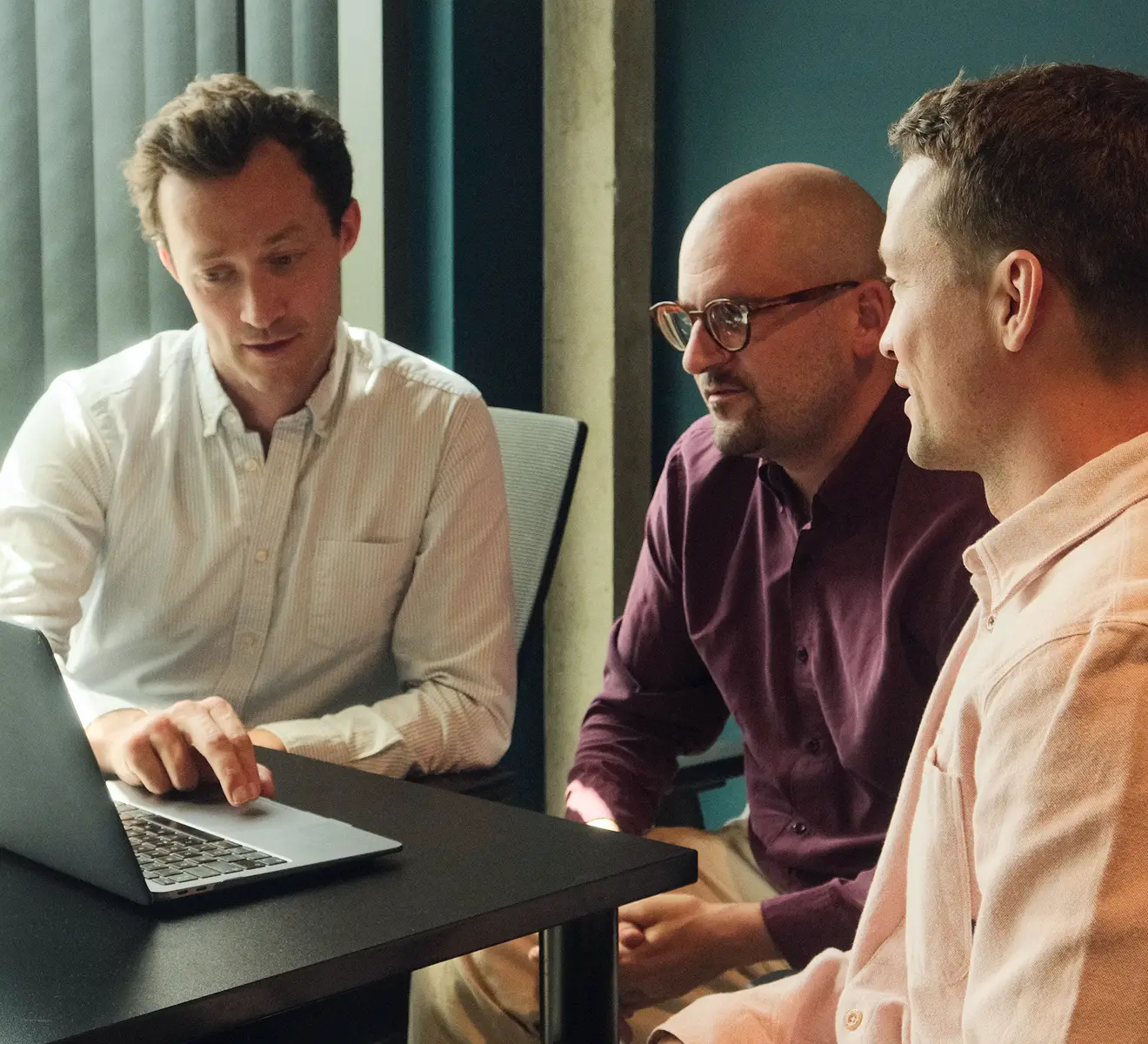 Three men sitting at a table engaged in discussion, with one man pointing at a laptop screen.