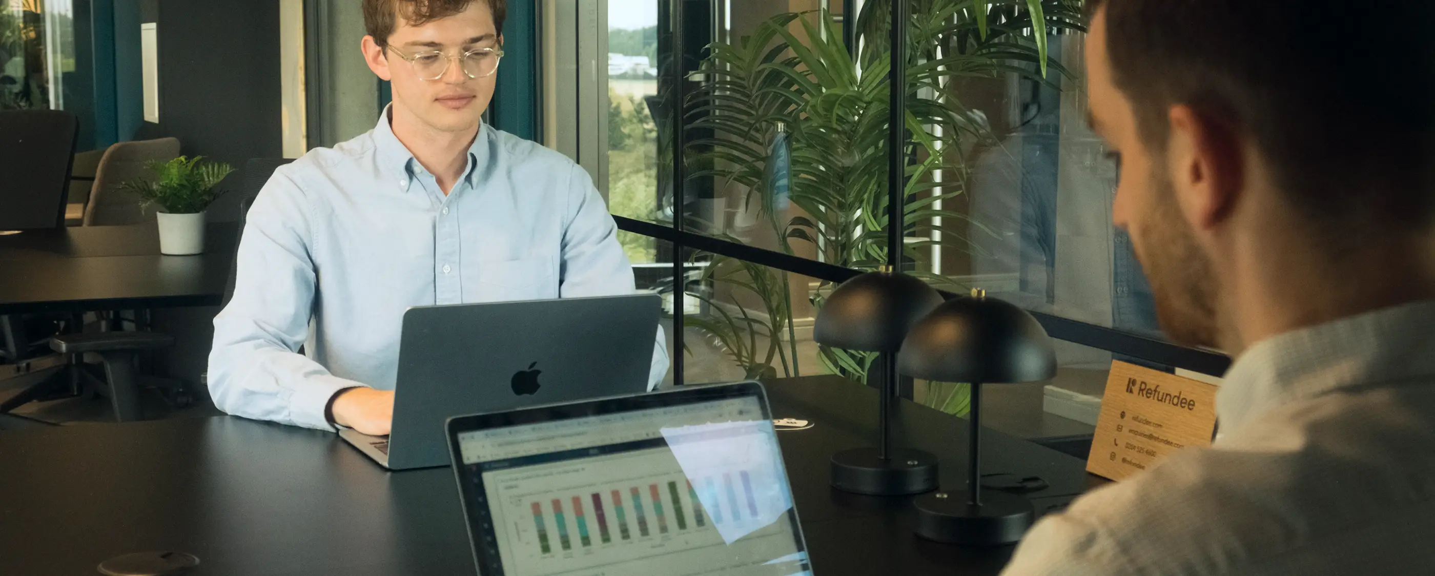Two men in an office working on laptops at a black table with plants and a sign labeled 'Refundee' in the background.