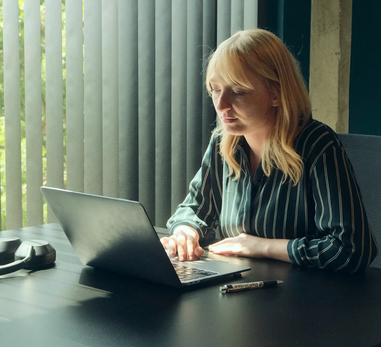 A woman with blonde hair wearing a dark green and white striped shirt is working on a laptop at a black table near a window with vertical blinds and green foliage outside.