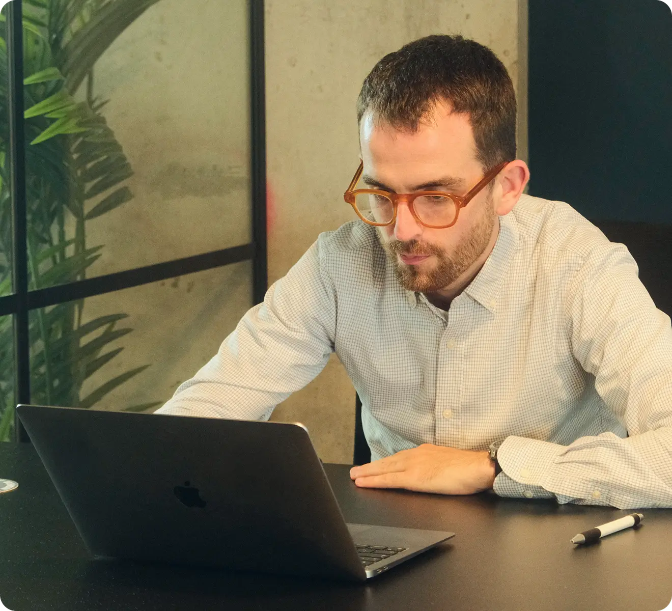 Man wearing glasses and a checked shirt, sitting at a desk and working on a laptop in a modern office with a plant in the background.