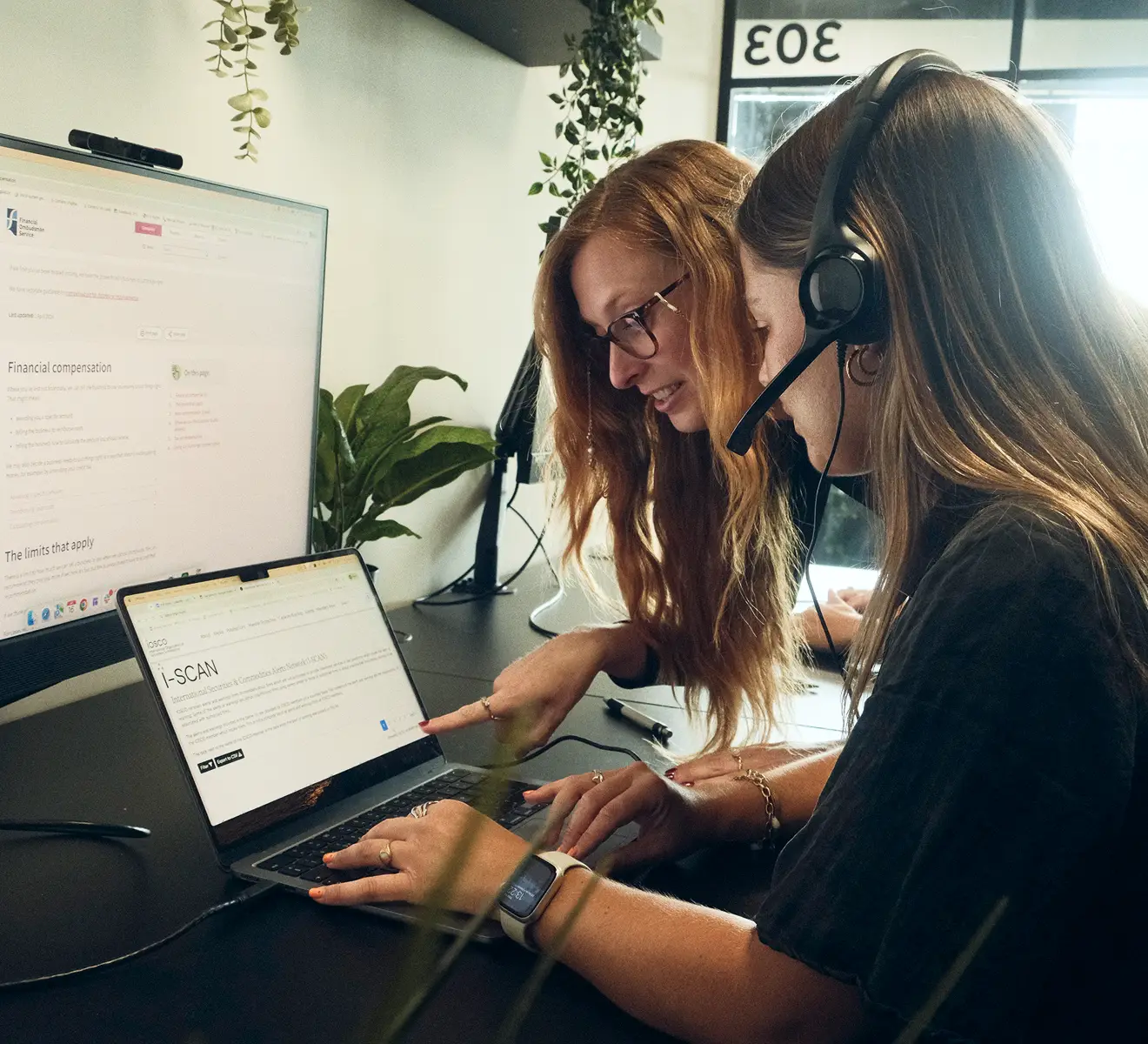 Two women working together at a desk with a laptop and a large monitor; one woman with long red hair and glasses points at the laptop screen while the other with a headset types, with green plants in the background.