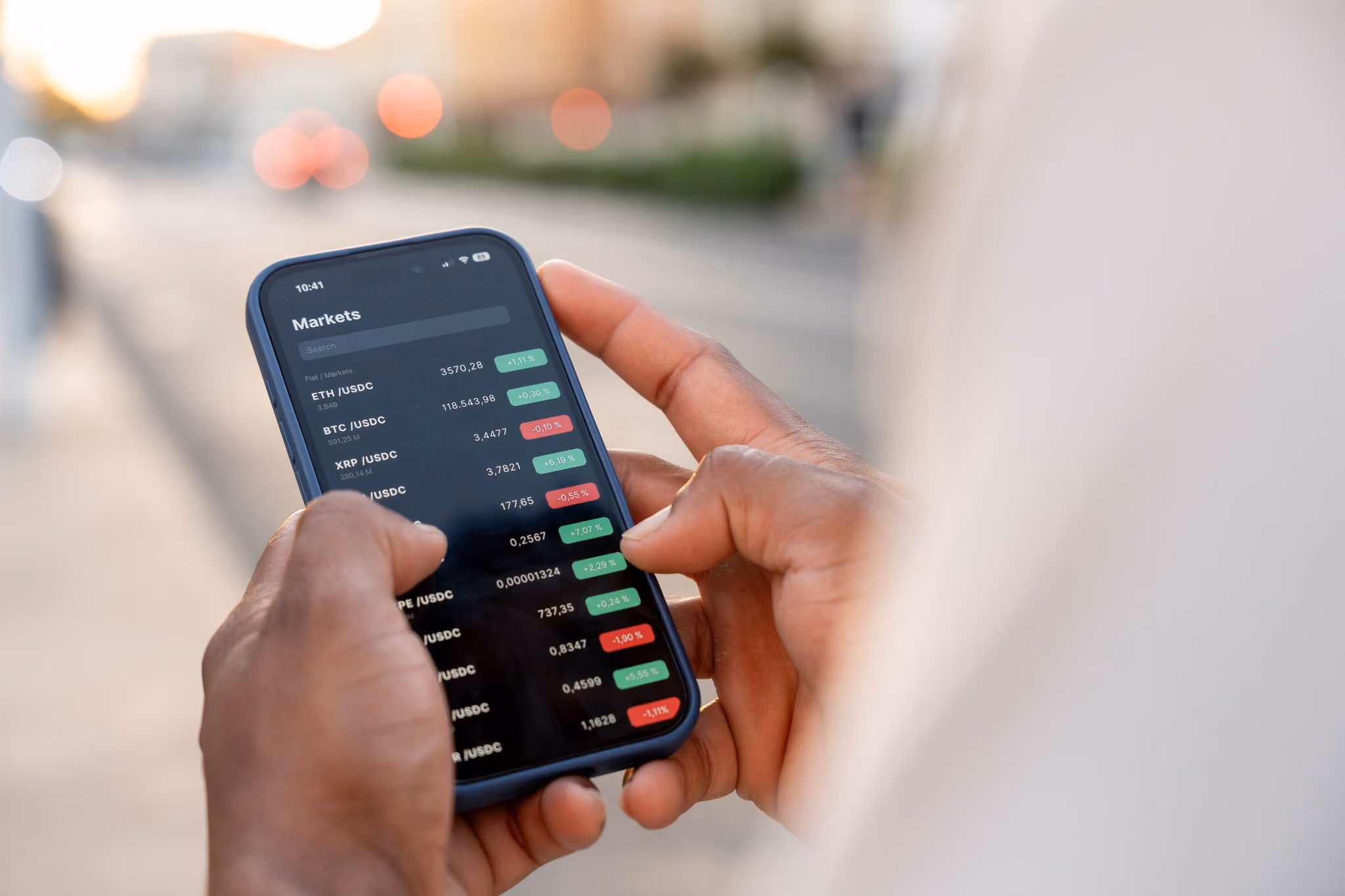 Close-up of hands holding a smartphone displaying a cryptocurrency market app with various crypto prices and percentage changes in green and red.