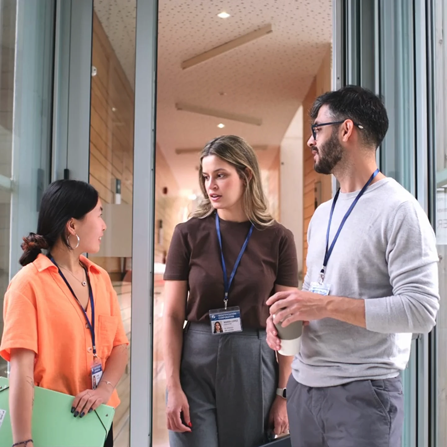 Three colleagues wearing ID badges standing and talking in a modern office hallway, one holding a green folder, another holding a coffee cup.