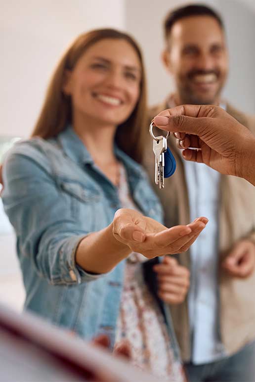 Smiling woman with open hand receiving house keys from a person, man stands smiling in background.