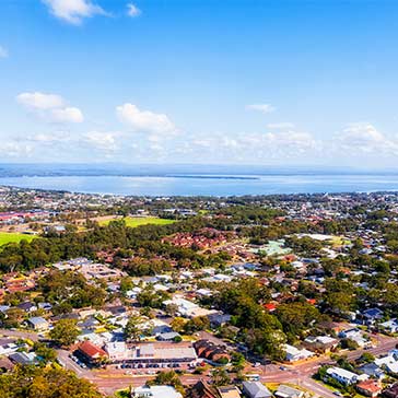 Aerial view of a coastal town with houses, trees, and water under a blue sky.