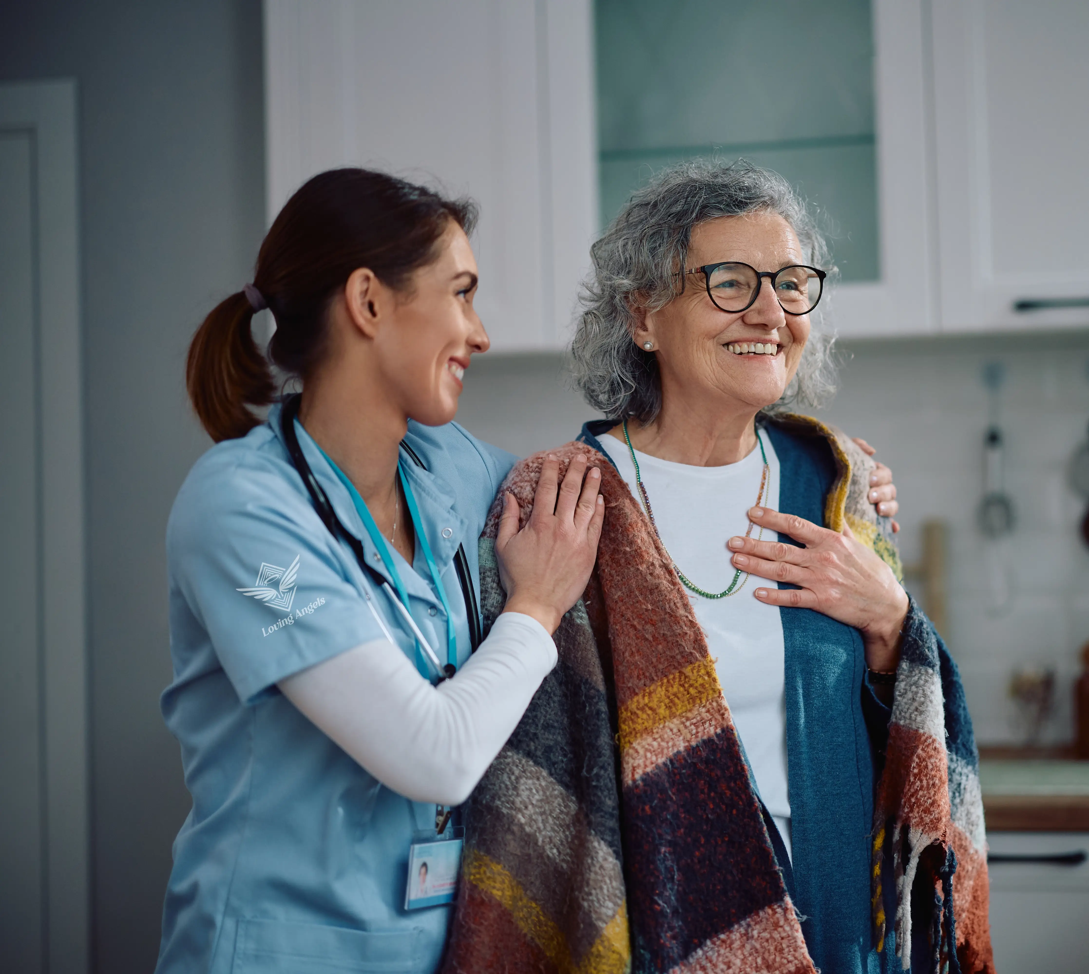 Caregiver smiling while wrapping a colorful blanket around a happy elderly woman with glasses.