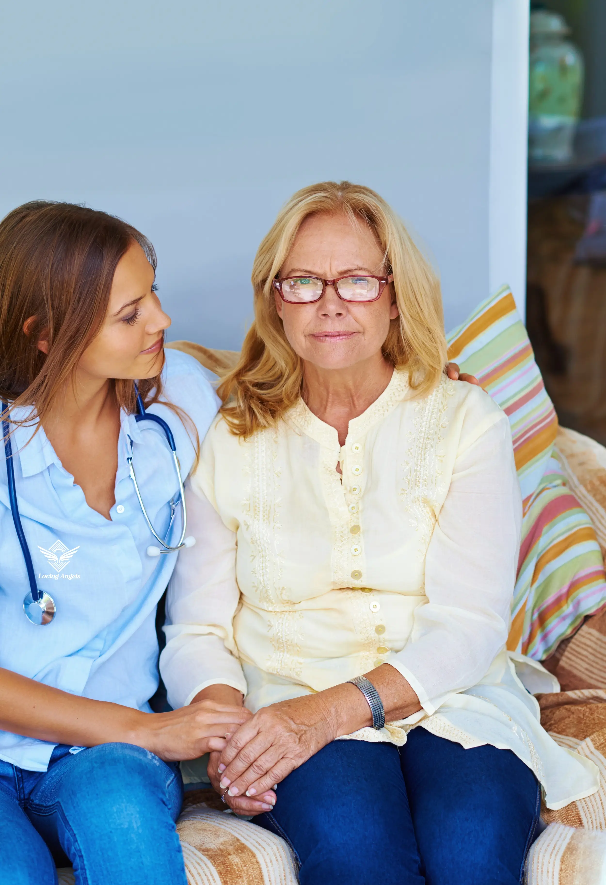 Young female doctor comforting older woman sitting on couch holding hands.