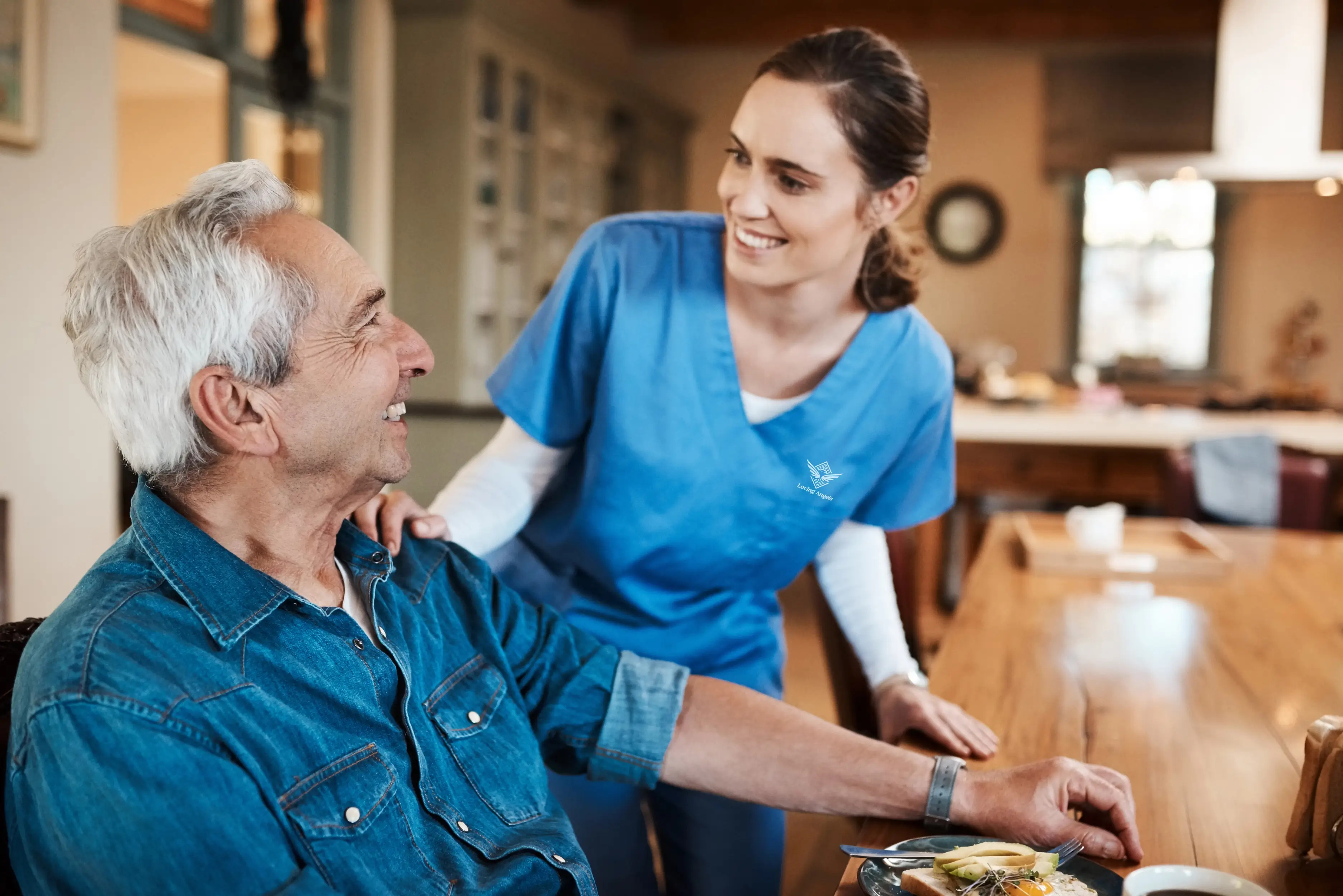 Smiling caregiver in blue scrubs comforting a seated elderly man during breakfast.